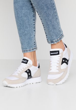 Person trägt hellblaue Jeans und weiße Saucony-Sneaker mit schwarzen Akzenten und steht auf einem hellgrauen Boden.