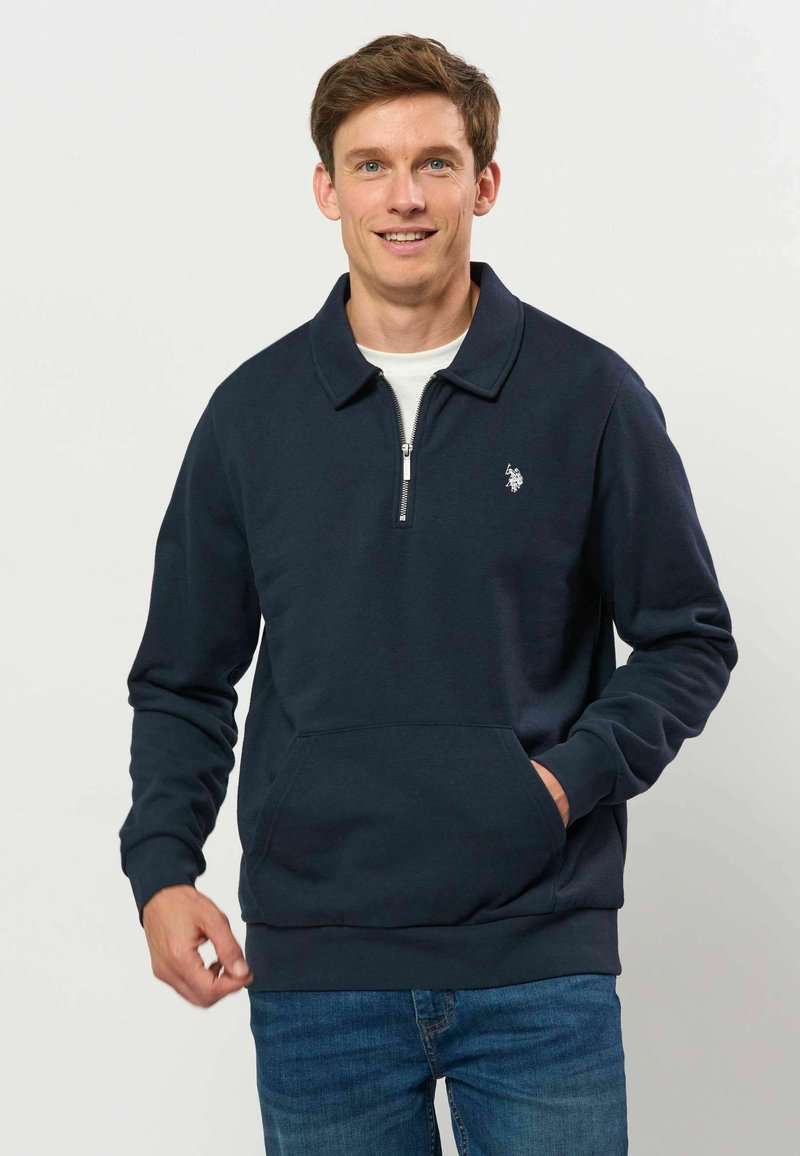 Jeune homme aux cheveux bruns courts souriant, portant un pull bleu marine foncé avec fermeture éclair au quart et un jean bleu sur un fond clair uni.