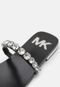 Sandale plate noire avec de grands strass transparents sur la bride et le logo "MK" sur la semelle, sur un fond blanc.