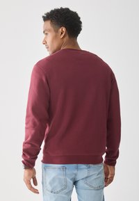 Man with short curly hair wearing a burgundy long-sleeve shirt and light blue jeans, facing away against a plain white background.