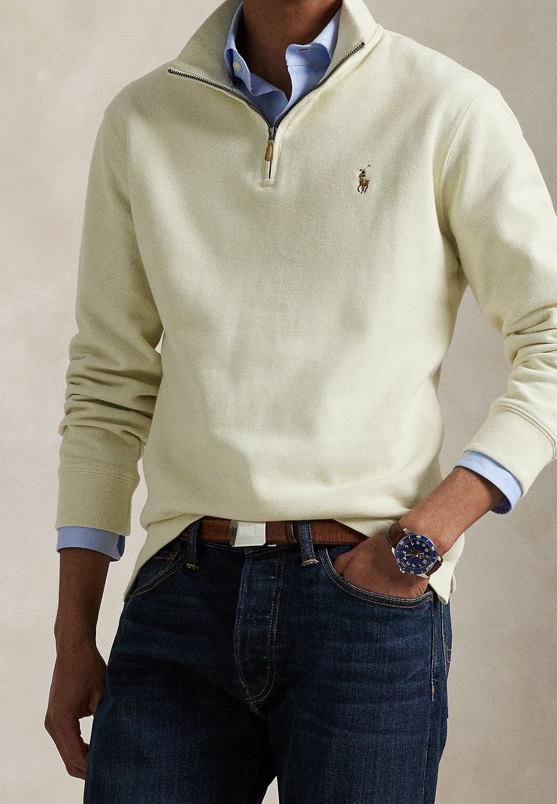 Homme portant un pull crème à zip quart sur une chemise bleu clair, jean foncé, ceinture marron et montre-bracelet bleue, main dans la poche.
