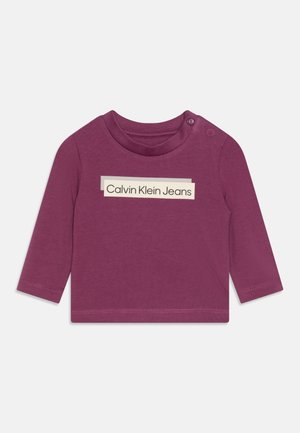 Fioletowa koszula z długim rękawem, z okrągłym dekoltem, zatrzaskami na ramieniu oraz prostokątnym logo "Calvin Klein Jeans" w kremowym kolorze.