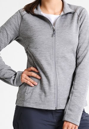 Femme portant une veste zippée intégrale gris clair avec col montant, main sur la hanche, et pantalon foncé sur fond blanc.