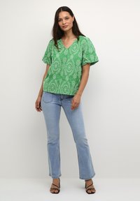 Zāles zaļas kokvilnas maisījuma tops ar baltajiem paisleja rakstiem, V veida izgriezums un īsām pūkainām piedurknēm, apvienots ar gaiši ziliem platām biksēm un melnām sandālēm.