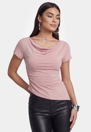 Frau trägt ein rosa Kurzarm-Top mit drapiertem Ausschnitt und schwarze Lederhose, dazu große Creolen und ein silbernes Armband.