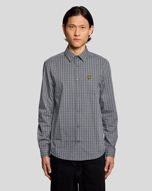 Bărbat purtând o cămașă cu mânecă lungă, în carouri alb-negru, cu un logo galben pe pieptul stâng, stând în fața unui fundal simplu, deschis la culoare.