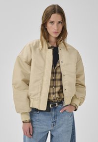 Beige Bomberjacke mit glänzendem Finish, gerippten Bündchen und Druckknopfverschluss, kombiniert mit einem karierten Hemd und Jeans.