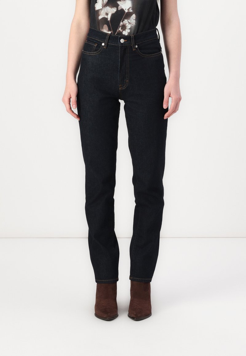 Jeans en denim foncé taille haute avec une coupe slim, présentant des coutures dorées, une fermeture à boutons à l'avant et des poches arrière.