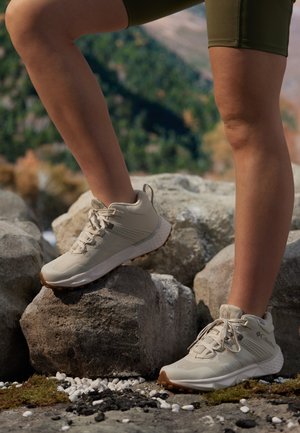 Beżowe buty sportowe z teksturowanej siateczki i syntetycznych materiałów, z zapięciem na sznurówki i gumową podeszwą, umieszczone na skalistym terenie.