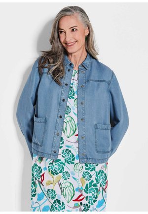 Jeansjacke in Hellblau, mit durchgehender Knopfleiste, zwei aufgesetzten Taschen und lockerer Passform. Über einem blumengemusterten Kleid getragen.