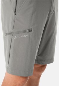 Pantalones cortos ligeros de color gris claro con un bolsillo con cremallera en el lateral, hechos de una tela suave y ligera. Presenta el logo de Vaude impreso cerca del bolsillo.