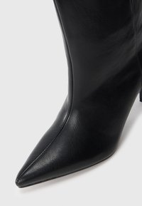 Primer plano de una bota de tacón alto negra de cuero con punta afilada y costuras visibles sobre un fondo claro y sencillo.