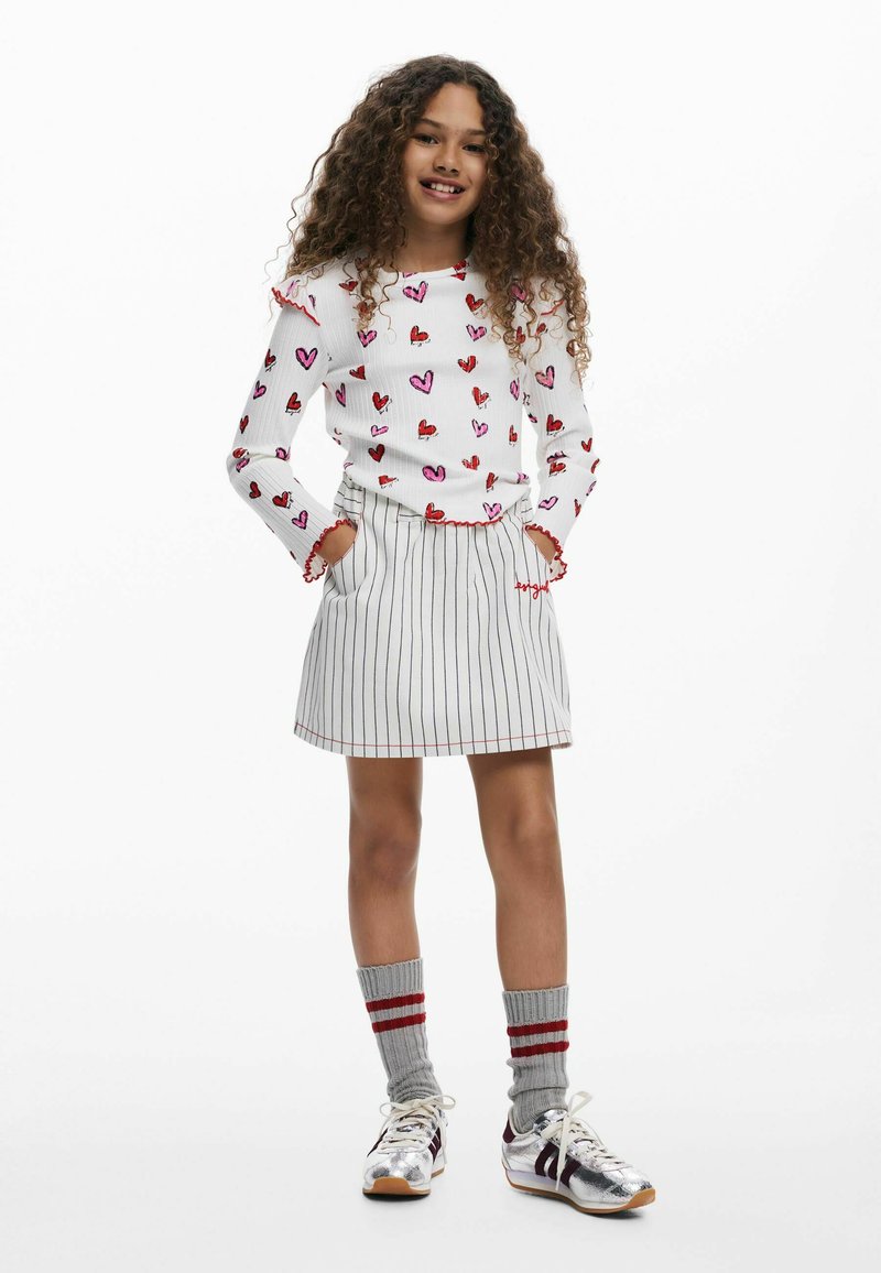 Weißes Langarmhemd mit Herzmustern, rotem Saum. Gestreifter Skort mit roten Akzenten. Graue Socken mit roten Streifen, silberne Turnschuhe.