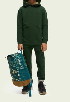 Sweat à capuche vert avec une poche avant, associé à un pantalon de jogging assorti. Tient un sac à dos turquoise et marron avec une sangle bleue.