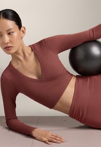 Donna con abbigliamento sportivo color ruggine che tiene una palla da ginnastica nera mentre è sdraiata di lato su un tappetino da yoga.