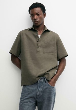 Mann trägt olivgrünes kurzärmeliges Hemd mit Brusttasche und blauer Jeans, steht vor schlichtem weißem Hintergrund.