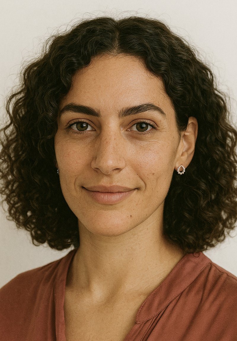 Porträtt av en person med krulligt mörkt hår, iklädd en top i rostfärg. De har subtila fräknar och är prydde med små, ovalformade örhängen.