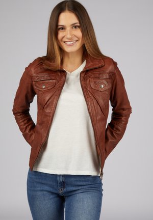 Veste en cuir marron avec fermeture éclair à l'avant, poches poitrine à boutons pression et col structuré. Portée par-dessus une chemise blanche et un jean bleu.