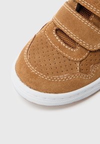 Baskets en daim marron avec semelle en caoutchouc blanche, présentant un design perforé, des accents cousus et des brides Velcro ajustables.