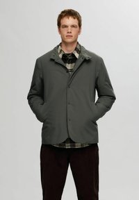 Grå, lett jakke med knapper foran, sidelommer og høy krage, lagd over en svart-og-hvit rutete skjorte.