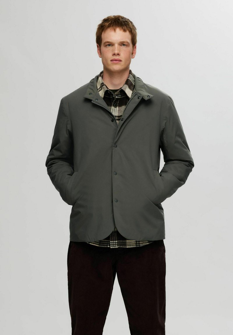 Grå, lett jakke med knapper foran, sidelommer og høy krage, lagd over en svart-og-hvit rutete skjorte.