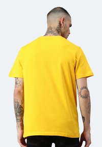Gelbes Baumwoll-T-Shirt mit kurzen Ärmeln, Rundhalsausschnitt und einer glatten Textur. Sichtbare, freiliegende Tattoos auf den Armen.