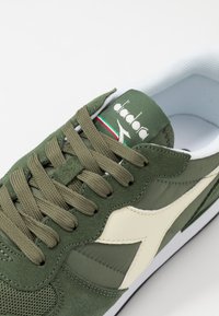 Green suede and fabric sneaker with beige accents, flat olive laces, perforated toe area, and branding on the tongue, set against a white background.