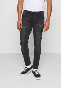 Jeans de mezclilla oscura con un corte ajustado, que presentan sutiles detalles desgastados y toques de pintura salpicada. Combinados con zapatillas negras y calcetines blancos.