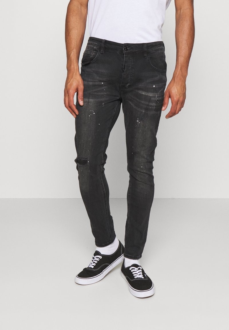 Jeans de mezclilla oscura con un corte ajustado, que presentan sutiles detalles desgastados y toques de pintura salpicada. Combinados con zapatillas negras y calcetines blancos.