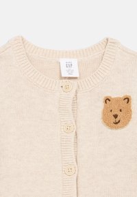 Krém színű baba kardigán három gombbal és egy kis, texturált mackóarc folttal a mellkason.