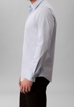 Homme portant une chemise bleu clair à manches longues boutonnée et un pantalon noir, debout de profil sur un fond gris.