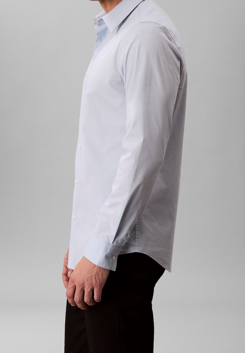 Homme portant une chemise bleu clair à manches longues boutonnée et un pantalon noir, debout de profil sur un fond gris.