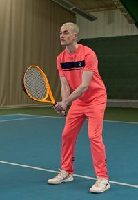 Světlý korálový sportovní outfit s krátkými rukávy a černými pruhy, doplněný bílými sportovními botami. Hráč drží tenisovou raketu.