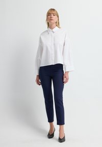Camisa blanca recortada con cuello, que presenta mangas largas y una textura delicada, combinada con pantalones ajustados azul marino y tacones negros.