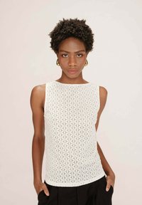 Haut sans manches blanc avec un motif de dentelle géométrique, texture lisse et encolure ronde, associé à un pantalon noir.