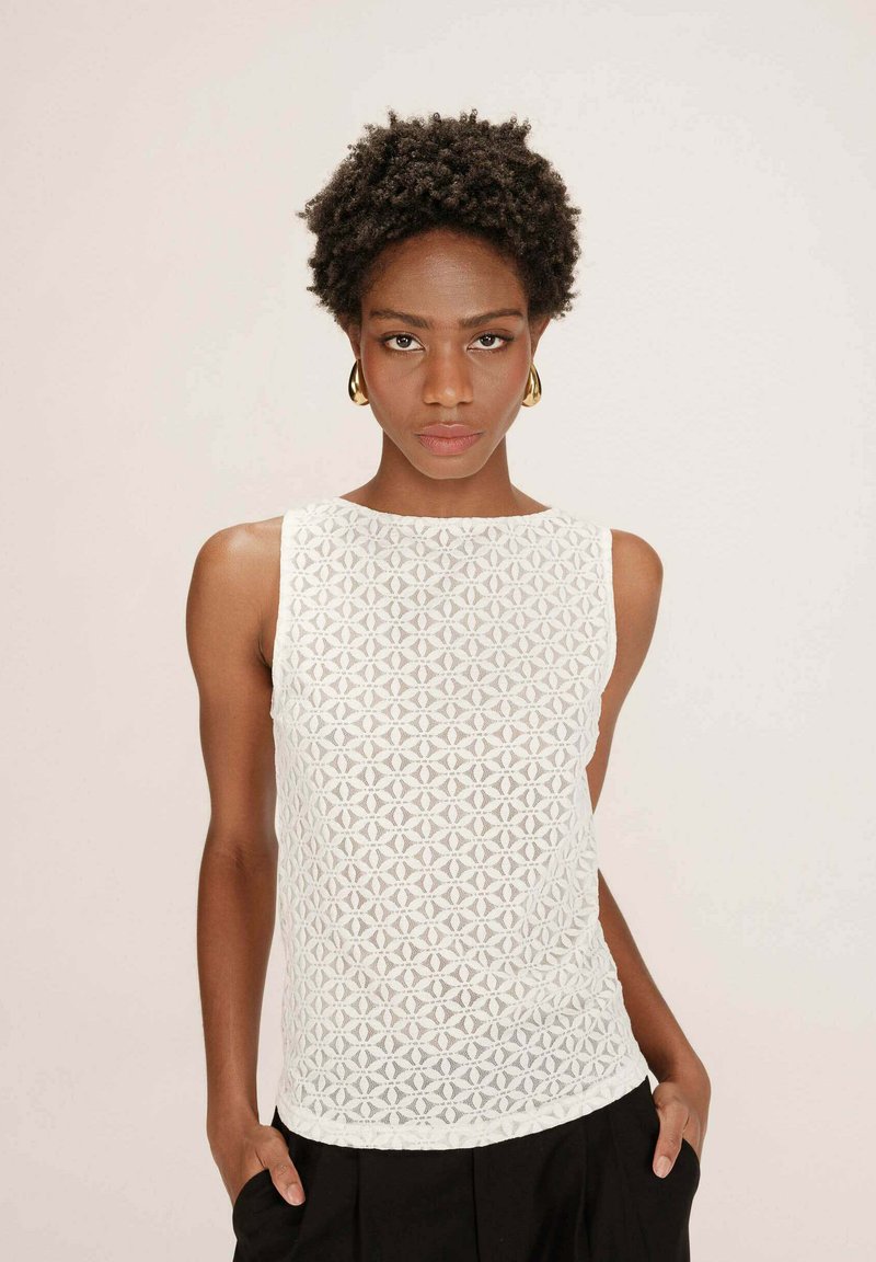 Haut sans manches blanc avec un motif de dentelle géométrique, texture lisse et encolure ronde, associé à un pantalon noir.