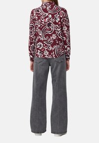 Vrouw in een bordeauxrode, lang mouwige bloempatronen shirt en hoge, loose-fit grijze jeans, staand met haar rug naar voren gericht.