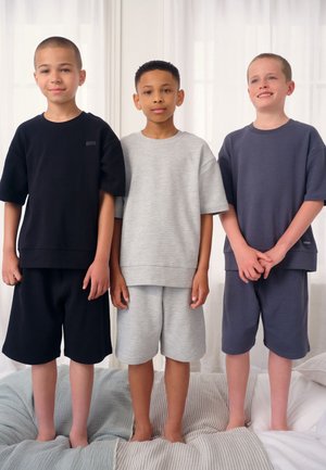 Trzech chłopców stoi boso na łóżku, ubrani w pasujące koszulki z krótkim rękawem i szorty w kolorze czarnym, szarym i ciemnoniebieskim.