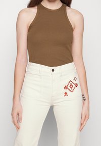 Kvinna som bär en brun ärmlös topp och högmidjade off-white jeans med röd geometrisk broderi på framlappen.