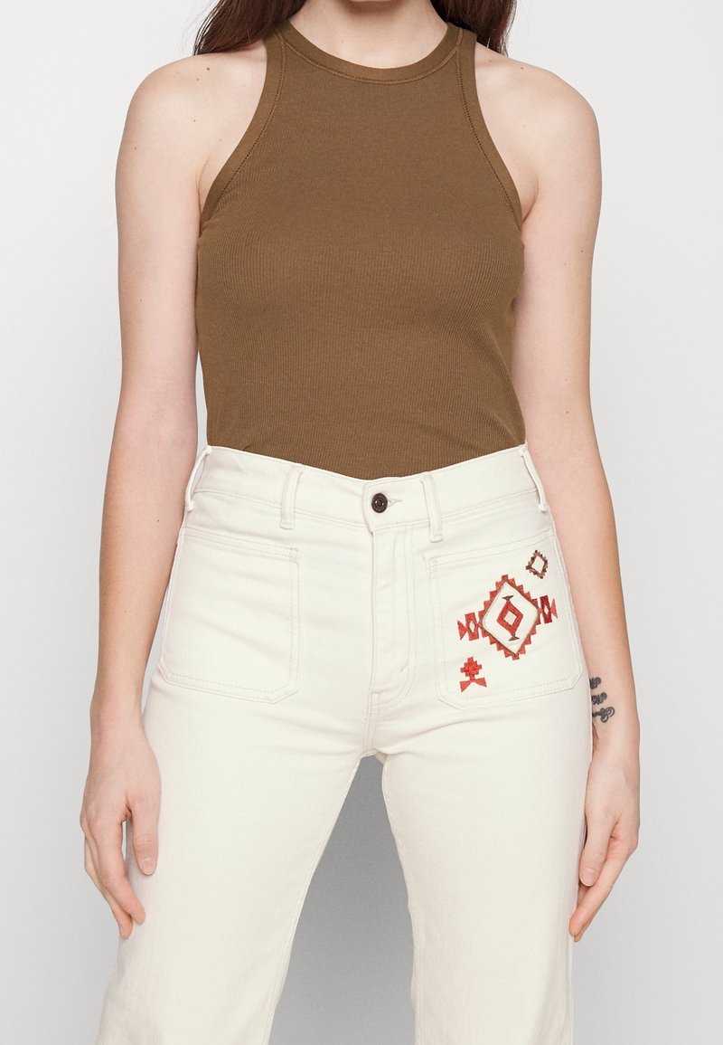 Kvinna som bär en brun ärmlös topp och högmidjade off-white jeans med röd geometrisk broderi på framlappen.