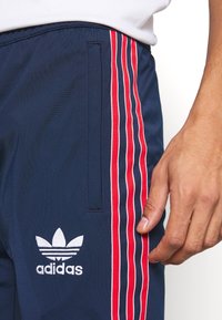 Tmavě modré sportovní kalhoty s třemi červenobílými svislými pruhy na boku a vyšívaným bílým logem Adidas na spodní části nohy.