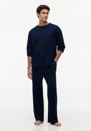 Jeune homme debout, pieds nus, portant une chemise à manches longues bleu marine et un pantalon large assorti sur un fond blanc uni.