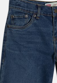 Jeans de mezclilla con un lavado azul oscuro. Presenta herrajes de cobre, cinco bolsillos y costuras doradas. El material parece suave con una ligera textura.