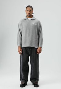 Lichtgrijze fleece trui met kraag en geborduurd logo, gecombineerd met losse zwarte jeans en zwarte laarzen met roze accenten.