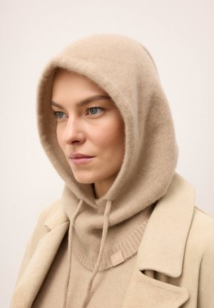 Mujer con suéter beige de punto con capucha y cordones, y un abrigo beige a juego, mirando ligeramente hacia un lado sobre un fondo liso.