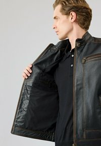 Zwarte leren jack met een gladde textuur, ritszakken en een bruine accent langs de naden. De binnenvoering is eenvoudig zwart.