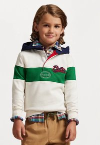 Balts sporta džemperis ar zaļām un tumši zila akcentiem, ar apkakli un izšūtu logotipu. Valkāts virs pleda krekla, kopā ar bēšām biksēm.