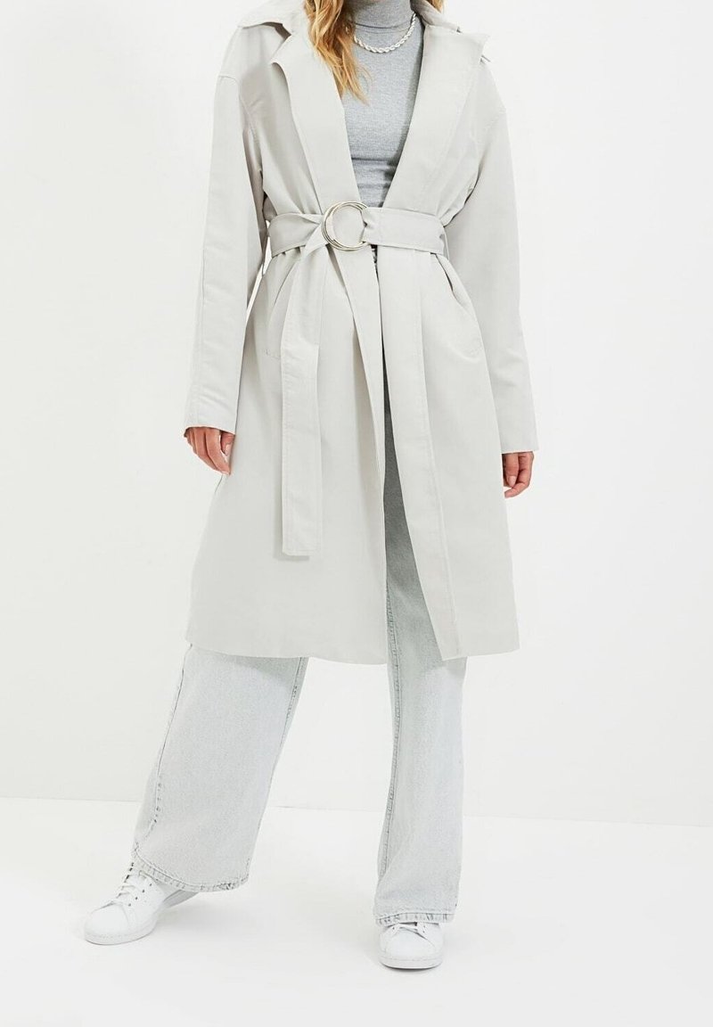 Abrigo trench de color beige claro con cinturón sobre un suéter de cuello alto gris, combinado con jeans de pierna ancha de color claro y zapatillas blancas sobre un fondo blanco liso.