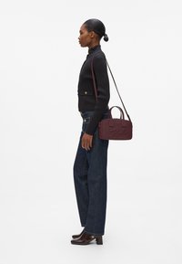 Sac à bandoulière bordeaux avec poignées supérieures, matériau texturé, logo embossé et bandoulière ajustable, associé à un jean foncé et un pull noir côtelé.