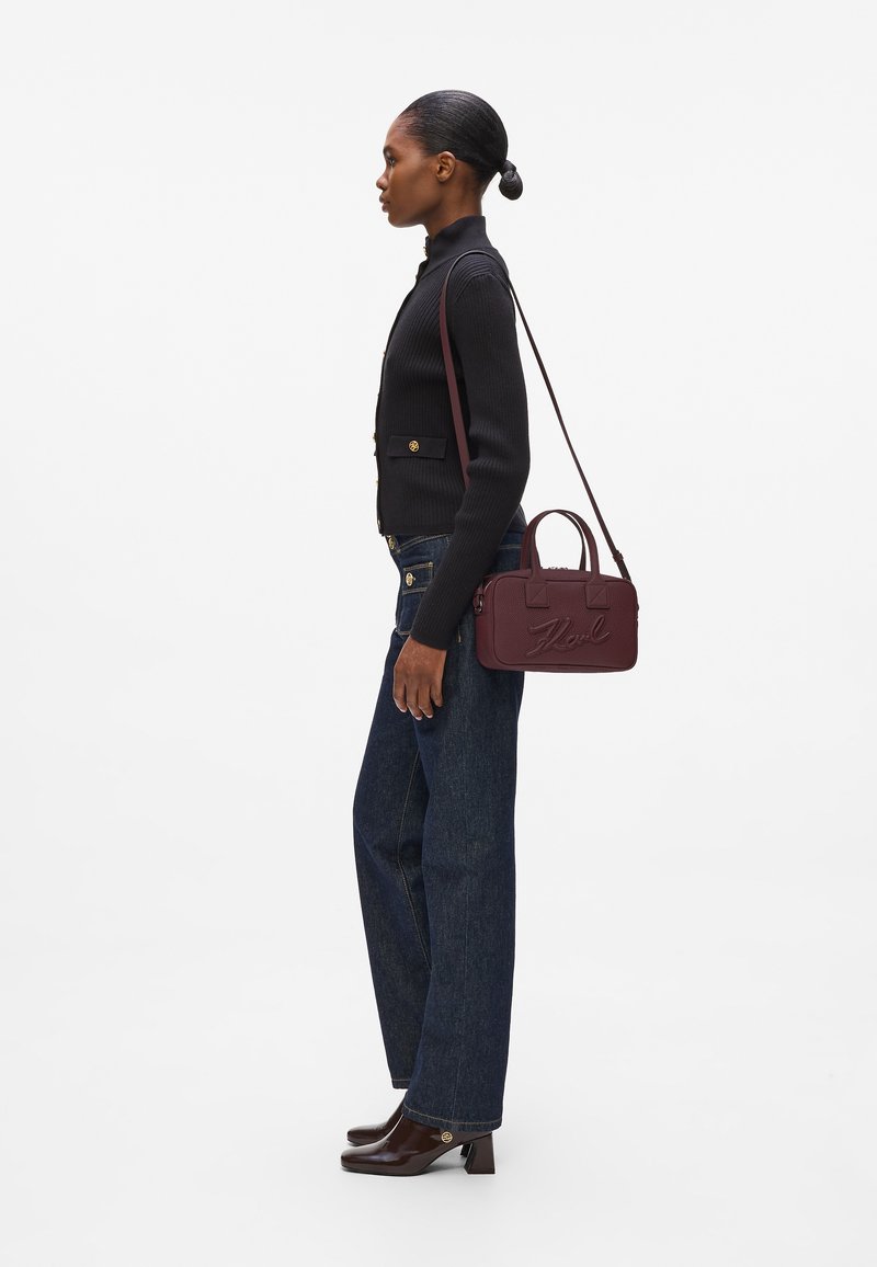 Sac à bandoulière bordeaux avec poignées supérieures, matériau texturé, logo embossé et bandoulière ajustable, associé à un jean foncé et un pull noir côtelé.